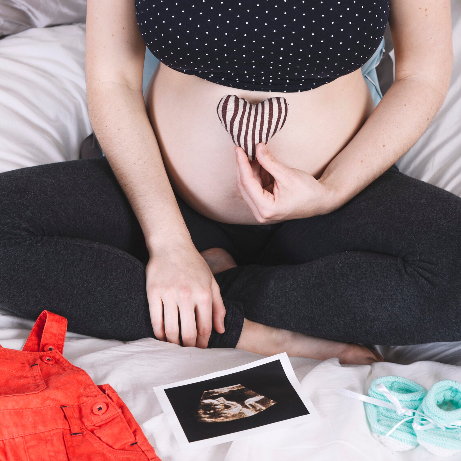 Pregnant woman sitting on a bed holding a fabric heart on her bare belly with an ultrasound photo, baby booties, and baby clothes beside her.