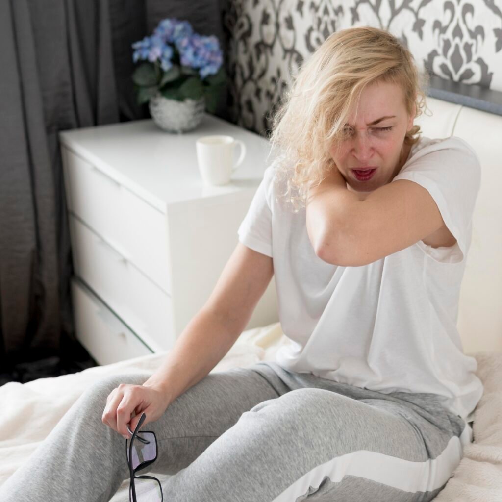 Woman sneezing into her elbow while sitting on bed, possibly indicating an underlying health condition like rapidly progressive renal failure.